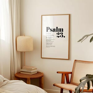 May include: A framed print with a white background and black text that reads "Psalm 23." The print is hanging on a white wall in a room with a wooden chair, a lamp, and a side table.