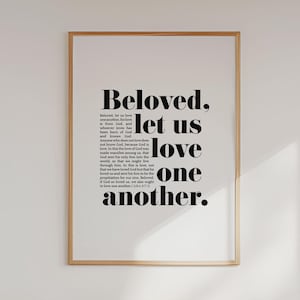 May include: A framed print with the words "Beloved, let us love one another" in a bold, black font. The print has a white background and a thin, light brown frame. The text is a quote from the Bible.