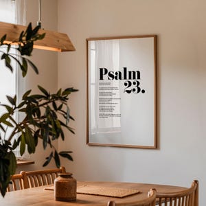 May include: A framed print with a white background and black text. The text reads "Psalm 23." The print is hanging on a white wall in a room with a wooden table and chairs.