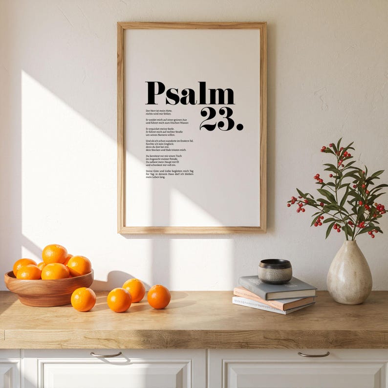 May include: A framed print with a white background and black text. The text reads "Psalm 23." and includes a poem in German. The print is hanging on a white wall above a wooden countertop with a bowl of oranges and a vase of red berries.