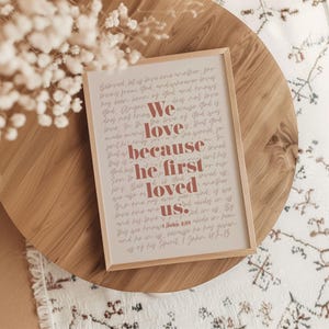 May include: A framed print with a light wood frame, featuring a quote from 1 John 4:19. The text is in a mix of fonts and colours, with the words "We love because he first loved us" highlighted in a reddish-brown hue.