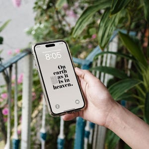 Peut inclure: Un smartphone avec une coque transparente affiche l'heure 8h05 et le texte "On earth as it is in heaven." Le t&eacute;l&eacute;phone est tenu devant un fond de verdure et une balustrade bleue et blanche.