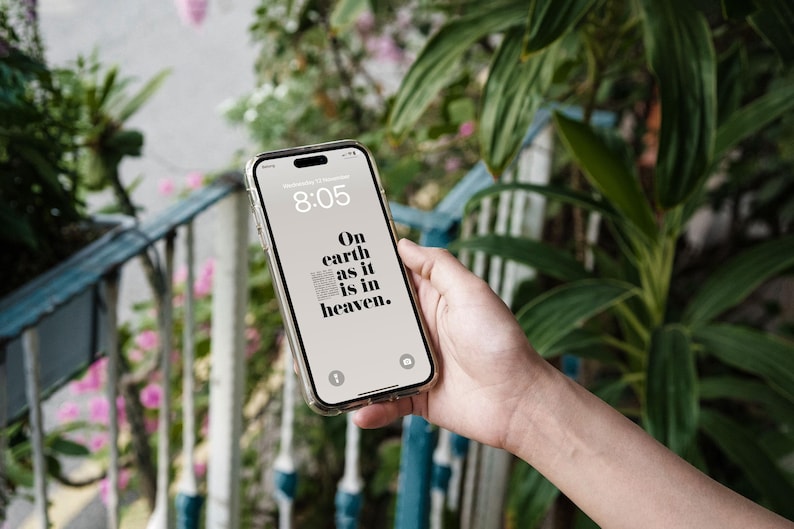 Peut inclure: Un smartphone avec une coque transparente affiche l'heure 8h05 et le texte "On earth as it is in heaven." Le t&eacute;l&eacute;phone est tenu devant un fond de verdure et une balustrade bleue et blanche.