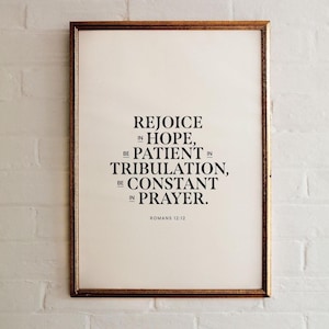 May include: A framed print with a gold-colored frame. The print features the text "REJOICE IN HOPE, BE PATIENT IN TRIBULATION, BE CONSTANT IN PRAYER. ROMANS 12:12" in a black, elegant font on a cream-colored background.