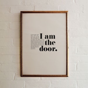 May include: A framed print with a white background and black text that reads "I am the door." The text is in a bold, sans-serif font. The frame is gold.