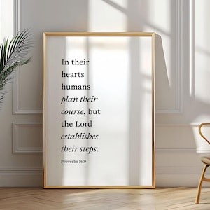 May include: A gold framed print with a white background and black text. The text reads "In their hearts humans plan their course, but the Lord establishes their steps. Proverbs 16:9".