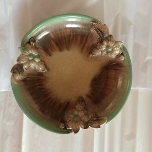 Peut inclure: Un bol en céramique vert et brun avec des accents floraux. Le bol a un bord festonné et un motif en relief au centre.