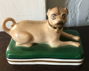 May include: A ceramic figurine of a pug dog wearing a gold collar and lying on a green, white, and gold striped cushion.