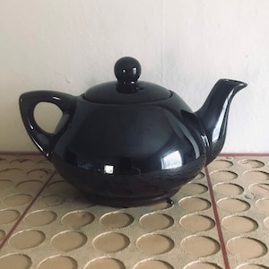 May include: A black ceramic teapot with a shiny finish and a rounded body. The teapot has a lid with a knob on top and a spout on one side.