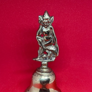 Op de afbeelding: Een kleine messing bel met een gedetailleerd beeldje van een zittend figuur met puntige oren en een ondeugende uitdrukking op zijn gezicht.