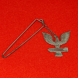 Peut inclure: Charme d'aigle argenté avec une main tendue, attaché à une chaîne argentée et une épingle de sûreté argentée.