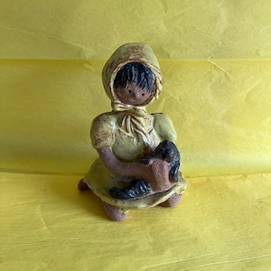Puede incluir: Una figurita de cerámica de una niña con un vestido amarillo y un gorro amarillo. Está sosteniendo un pequeño gato negro en sus brazos.