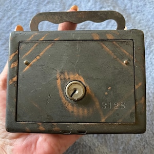 May include: A vintage metal money box with a keyhole and a handle. The box is painted black with a copper patina and has the number 3128 printed on the front.