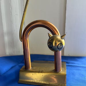 May include: A metal cat figurine featuring a copper-toned arched body, a brass-toned rectangular base, and a spherical head with pointed ears. A thin, straight brass rod extends upward from the back of the arch, representing a tail.