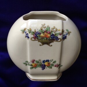 May include: A white ceramic vase with a round body and a square base. The vase is decorated with a floral design featuring a basket of fruit, including pears, grapes, and berries. The design is in shades of red, yellow, green, blue, and purple.