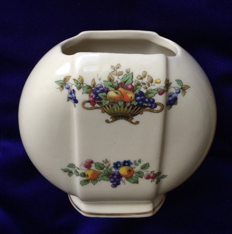 May include: A white ceramic vase with a round body and a square base. The vase is decorated with a floral design featuring a basket of fruit, including pears, grapes, and berries. The design is in shades of red, yellow, green, blue, and purple.