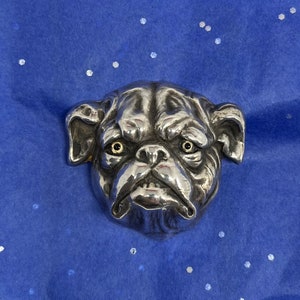 May include: A silver-colored bulldog head sculpture with detailed wrinkles and a slightly grumpy expression. The dog's eyes have a golden hue. The sculpture is set against a blue background with white speckles.