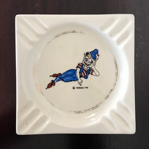 Peut inclure: Un cendrier carré en céramique blanche avec un lutin de dessin animé portant un chapeau bleu et un pantalon bleu. Le lutin est allongé et porte une chemise à rayures rouges et blanches. Le texte "WNAC-TV" est imprimé au bas du cendrier.