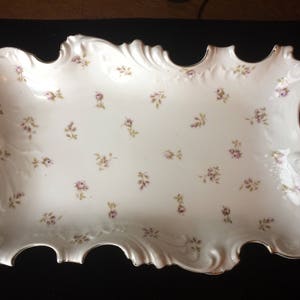 Op de afbeelding: Een wit porseleinen dienblad met een gegolfde rand en een delicaat bloemenpatroon. Het dienblad is versierd met kleine, roze bloemen en groene bladeren.