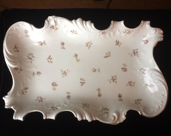May include: A white porcelain serving tray with a scalloped edge and a delicate floral pattern. The tray is decorated with small, pink flowers and green leaves.