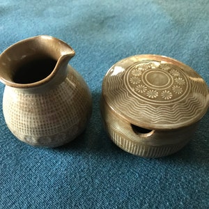 May include: A set of two brown ceramic containers with a decorative design. The larger container has a lid and a small opening on the side. The smaller container is a pitcher with a curved spout.