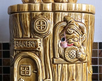 May include: A ceramic cookie jar shaped like a tree stump with a gnome on top. The gnome is holding a musical instrument and is wearing a red hat. The tree stump has a sign that says "BAKERY" and a window with another gnome inside holding a cookie.
