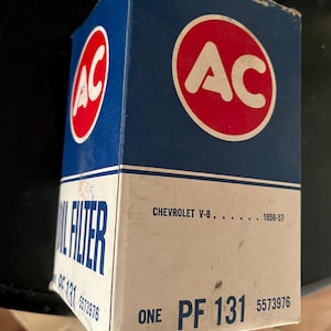 Peut inclure: Une boîte en carton bleu et blanc avec un cercle rouge contenant les lettres "AC" en blanc. La boîte est étiquetée "Filtre à huile PF 131" et "Chevrolet V-8 1956-57".
