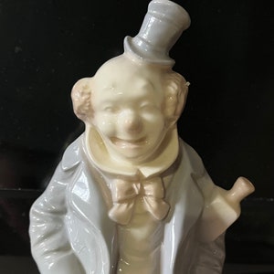 Peut inclure: Une figurine en porcelaine d'un clown portant un chapeau bleu et blanc, une veste bleue et un nœud papillon rose. Le clown tient une bouteille dans sa main droite.