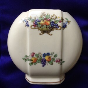 May include: A white ceramic vase with a rounded shape and a gold rim. The vase features a floral design with a basket of fruit and grapes.