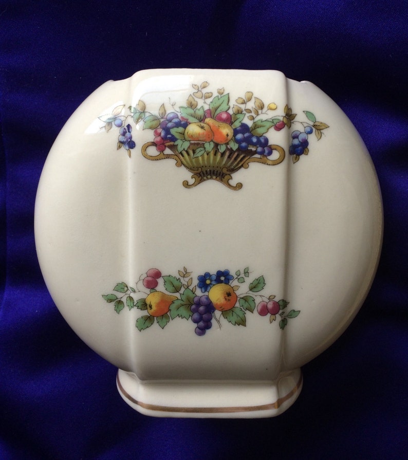 May include: A white ceramic vase with a rounded shape and a gold rim. The vase features a floral design with a basket of fruit and grapes.