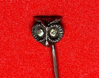 May include: A silver-toned owl-shaped hat pin with clear rhinestone eyes. The owl has detailed feathering and is set against a vibrant red background. The pin is a classic accessory.