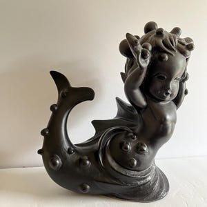 May include: A dark bronze-colored sculpture of a cherub with a mermaid tail. The cherub has its hands raised above its head, and the tail features a fin and decorative spheres. The sculpture is a decorative art piece.