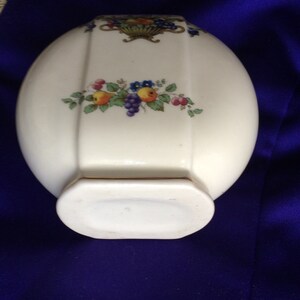 May include: A white ceramic vase with a gold trim and a floral design featuring red, yellow, and blue flowers and green leaves. The vase has a rounded oval shape with a flat bottom.