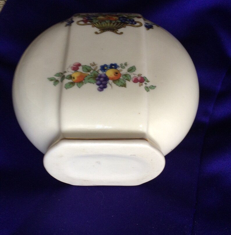May include: A white ceramic vase with a gold trim and a floral design featuring red, yellow, and blue flowers and green leaves. The vase has a rounded oval shape with a flat bottom.