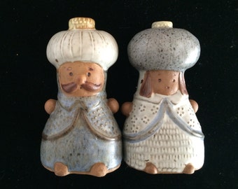 May include: A pair of ceramic salt and pepper shakers shaped like people wearing hats. The salt shaker is wearing a blue and white robe and has a brown beard and mustache. The pepper shaker is wearing a white and gray robe and has brown hair.