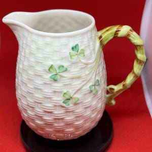 Puede incluir: Una jarra de crema de cerámica blanca con un patrón tejido y diseños de trébol verde. El asa es de diseño retorcido marrón y verde.