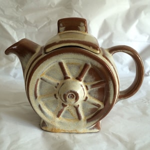 Puede incluir: Una tetera de cerámica marrón y blanca con forma de rueda. La tetera tiene una tapa y un asa.