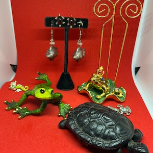 Pode incluir: Uma coleção de estatuetas decorativas, incluindo um sapo verde, uma tartaruga preta e um sapo dourado usando uma coroa, sentado em uma folha verde. As estatuetas são exibidas em uma superfície vermelha com um par de brincos de peixe de prata com pedras preciosas vermelhas.