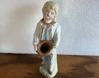 Puede incluir: Una figurita de porcelana de un niño pequeño con un atuendo blanco y azul que sostiene un sombrero marrón. La figurita está de pie sobre una base con un diseño floral.
