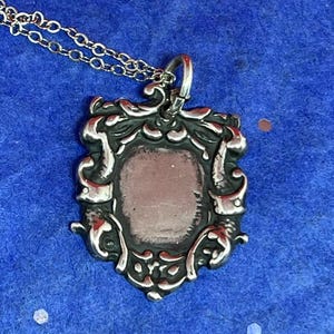 May include: A silver pendant necklace with a detailed ornate design. The pendant features a blank oval space in the center.