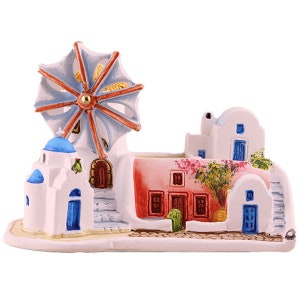 Könnte beinhalten: Eine weiße Keramikfigur, die eine griechische Dorfszene mit einer Windmühle, Häusern und einem Baum darstellt. Die Häuser sind in Weiß-, Blau- und Rosatönen bemalt, und die Windmühle hat blaue und rote Flügel.