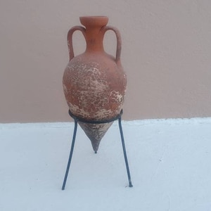 Peut inclure: Une amphore en terre cuite avec une base pointue et deux anses latérales, présentant une surface texturée et patinée avec des dépôts minéraux clairs. Le récipient est soutenu par un support tripode en métal noir à trois pieds fins.
