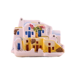 Può includere: Un modello in miniatura di un complesso di edifici in stile mediterraneo con pareti bianche, livelli inferiori gialli e porte blu. La struttura include piccole finestre, un tetto strutturato e una vite fiorita rosa dipinta sulla facciata.