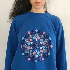 May include: A blue sweatshirt with a large, colorful patchwork design in the center. The design is a circular pattern of various shades of blue, pink, and white fabric.