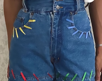 May include: High-waisted denim shorts with colorful ribbon detailing around the pockets and hem. The shorts are a light blue wash and have a relaxed fit.