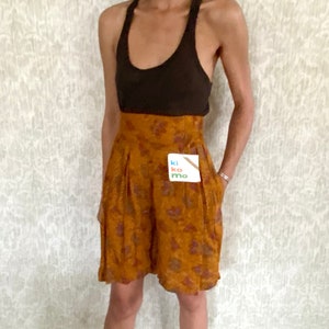 May include: A woman wearing a brown tank top and high-waisted, orange floral print shorts. She is wearing brown leather clogs. The shorts have a high waist and a wide leg.