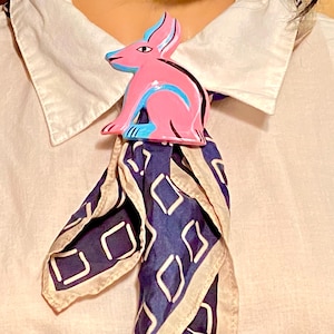 May include: A pink rabbit-shaped brooch with blue and black accents, worn on a patterned scarf. The scarf features a navy blue background with white diamond shapes. The brooch is attached to a white collared shirt.