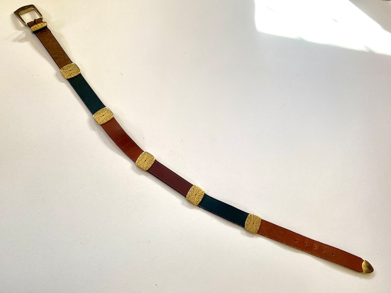 May include: A brown leather belt with a gold buckle and decorative metal accents. The belt has alternating sections of brown and black leather.