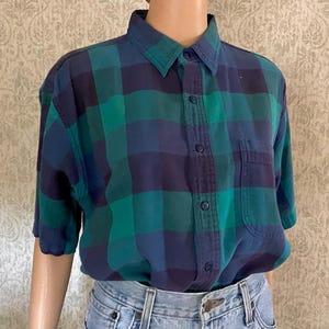 May include: A short-sleeved button-down shirt in a blue, green, and navy plaid pattern. The shirt has a collared neckline, a single chest pocket, and is worn with blue jeans. The shirt is a casual style.