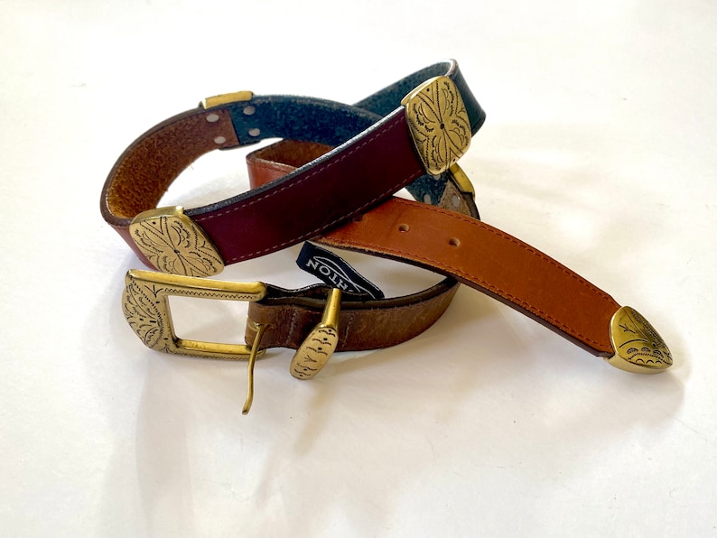 May include: Two leather belts with ornate brass buckles. The belts are brown, tan, and dark blue. The buckles have a floral design.